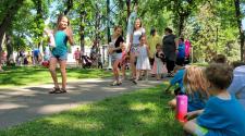 Kids and adults walking through Island Park with their stuffed animals for the Teddy Bear Parade while other families sit on the grass and watch