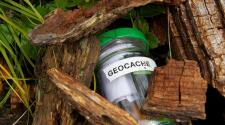 a jar with a green lid, jar labeled GEOCACHE with items in jar, semi-barried under wood