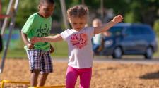 Little girl and little boy walking over little hurdles in park with boy swinging in background