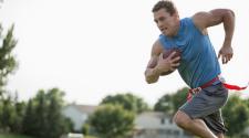 Man in the act of running with football in arm outside in a park playing flag football