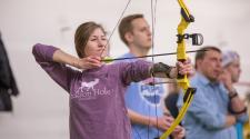 Woman shooting a bow and arrow with men in the background in a building