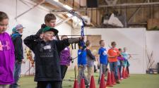 Young boy pulling back bow to shoot arrow with instructor standing behind and many kids lined up to shoot their arrow
