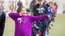 Young girl lining up bow and arrow to shoot with many other kids in the background lined up working on getting their bow and arrow ready to shoot indoors at archery lessons