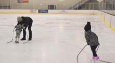 two young kids ice skating indoors at the Cornerstone Bank Arena using metal skate trainers with a male adult assisting one child skate