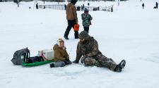 A boy and man sitting on the frozen pond next to a drilled ice fishing hole talking. The boy has a fishing pole in his hand and a sled full of ice fishing equipment sits behind the boy. In the background a boy and adult man walk . 