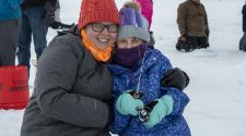 Woman and girl hug while smiling at the camera, all bundled up kneeling besides an ice fishing hole on a frozen pond. The girl holds a ice fishing rod. 