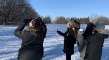 4 people bundled up in winter gear standing outside on a snow path in a field looking up at trees. One woman and a moan with binoculars up at their eyes looking through them. 