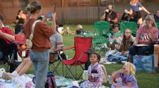 A woman stands and takes a picture of the two young girls sitting on a blanket in their PJs waiting on the lawn at Rheault Farm for the Movie in the Park movie to start with 20+ people in the background