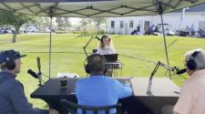 Female sits with a sound board and laptop in front of her managing the podcast of three men sitting in the foreground with head phones and mics.  This photo is set on the driving range tee box area of Osgood Golf Course on a sunny morning