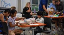 A young boy and a young girl sitting at a table with an orange table cloth doing crafts in a room full of people. 