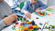Two kids sitting in chairs at a white table building with legos. 