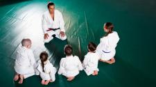 4 youth and instructor in Jiu Jitsu robes sitting on mat with their backs to camera in front of a male instructor  