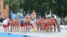 Large Group of Men and Women Lifeguards standing in a group around a few men lifeguards standing on a white tall lifeguard chair in front of the new Island Park Pool 