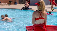 Woman lifeguard standing in front of Southwest Pool watching kids swim with a woman sitting on the side of the pool in the distance