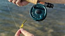 Close-up of hands holding a fly fishing rod and reel while tying a yellow-and-black fly lure, with water visible below.