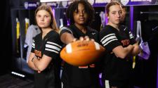 Three high school girls in black football uniforms stand in a locker room, one holding a football toward the camera.