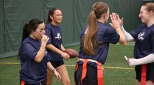 Four high school girls in practice shirts and flag belts smile and high-five during a flag football practice indoors.