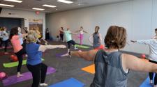 Older adults participate in a group fitness class, practicing balance and strength exercises on yoga mats in a community room.