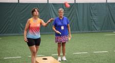 This image shows two women playing bean bag toss indoors on a turf field during the North Dakota Senior Games.