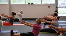 Adults participate in a group fitness class, seated on individual exercise mats with their legs extended while reaching their arms forward during a stretching and core exercise.