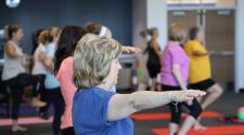 Adults participate in a group balance and fitness class, standing on individual mats while extending their arms and practicing balance poses.
