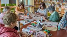 A group of older adults gathers around a table in a library for a craft activity, working with newspapers, scissors, glue and other materials.