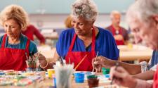 Older adults wearing red aprons participate in a group painting activity, using brushes and colorful paints around a shared table.
