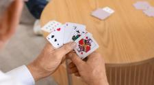Close-up of a person holding a hand of playing cards over a round wooden table, with additional cards on the table in the background.